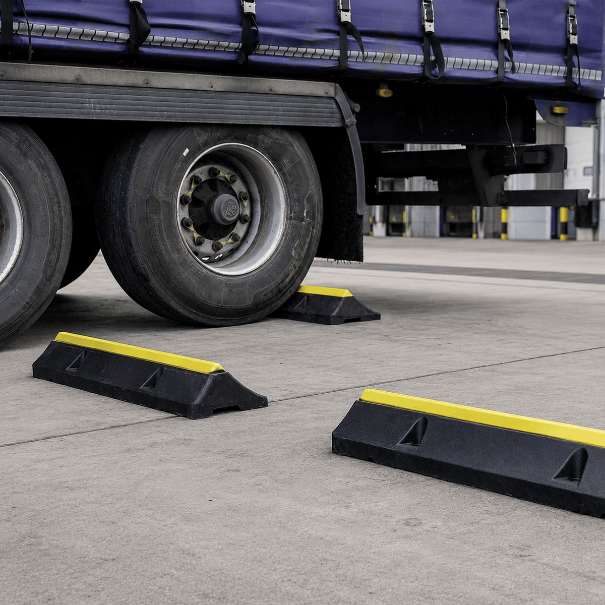 Black plastic truck wheel stoppers with bright yellow reflective stripes, shown placed on the concrete ground beside a semi-trailer tire at a truck stop.