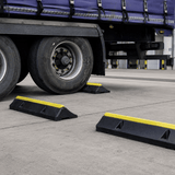 Black plastic truck wheel stoppers with bright yellow reflective stripes, shown placed on the concrete ground beside a semi-trailer tire at a truck stop.