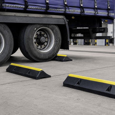 Black plastic truck wheel stoppers with bright yellow reflective stripes, shown placed on the concrete ground beside a semi-trailer tire at a truck stop.