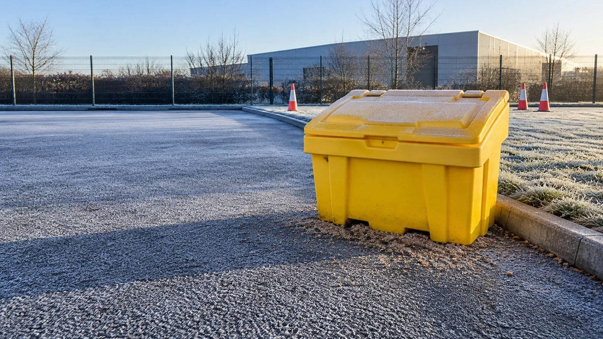 A yellow grit bin beside a kerb on a frost-covered commercial yard on a bright winter morning, traffic cones standing behind it